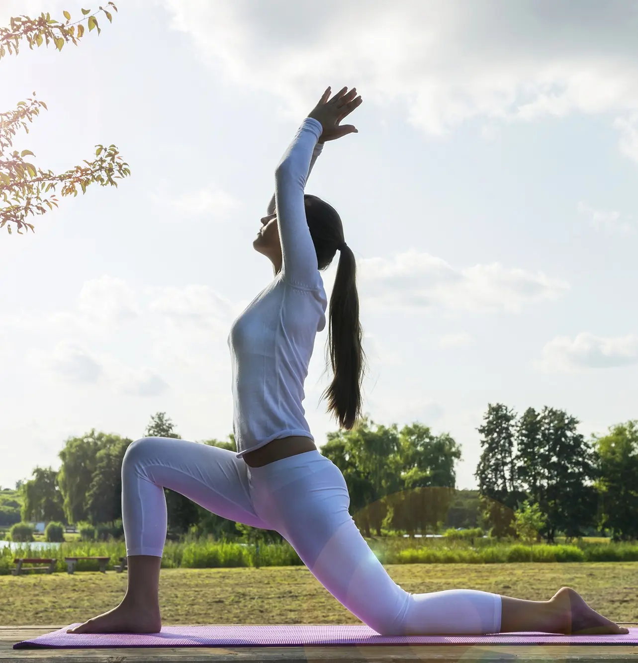 A woman in white yoga attire performs a lunge pose on a mat outdoors, with trees and a cloudy sky in the background, embodying wellness and tranquility.