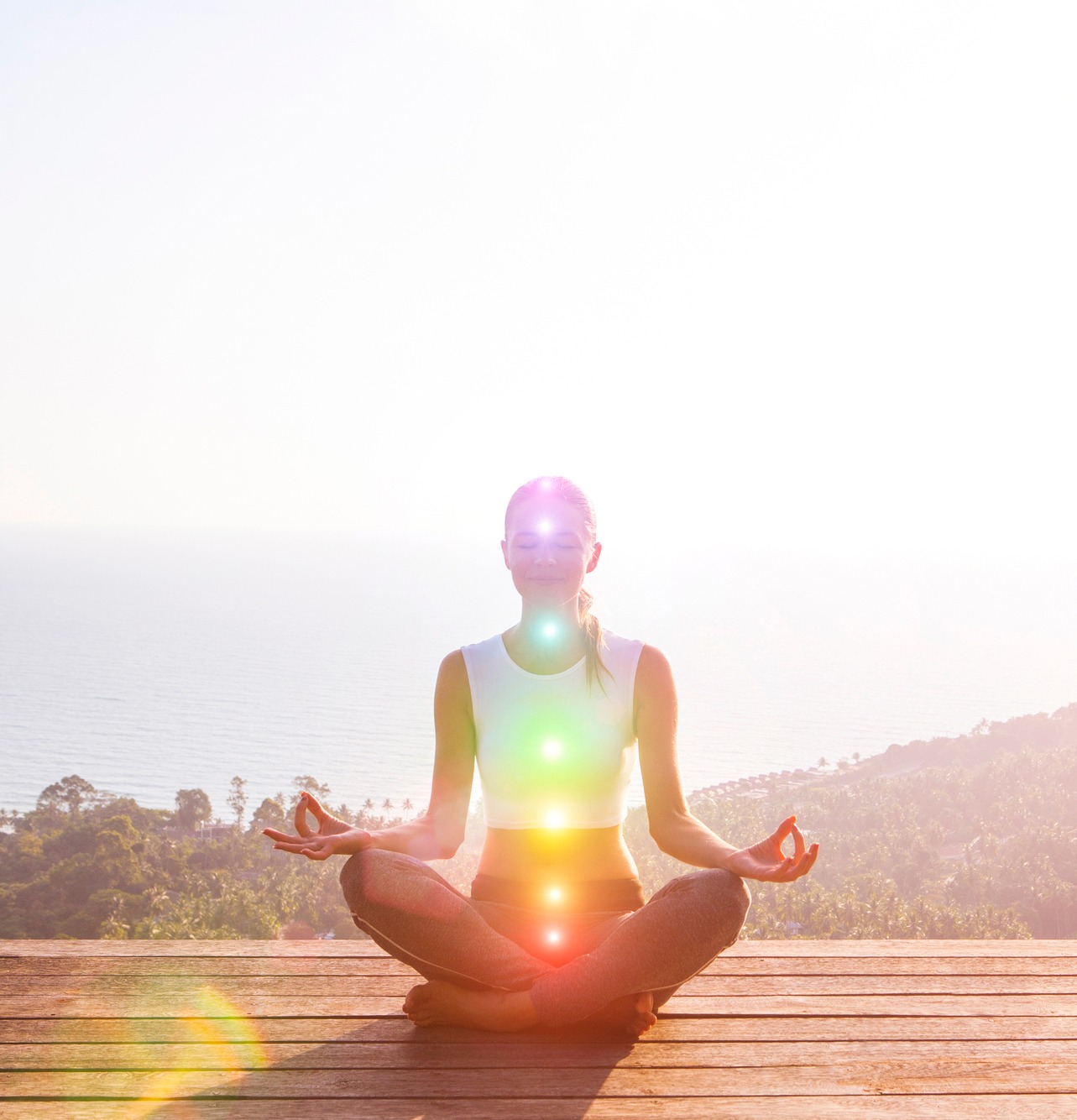 A woman sits in a meditative pose on a wooden deck overlooking the ocean, with vibrant energy points glowing along her torso.
