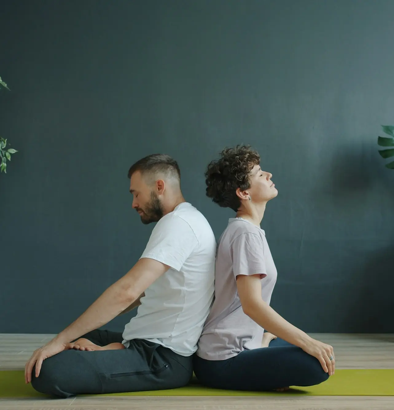 Couple seated back-to-back on a yoga mat, practicing mindfulness together in a serene indoor setting. Focus on harmony and connection.